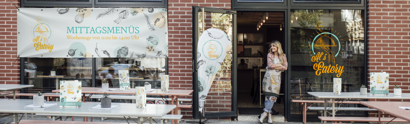 Eine Frau in einer Schürze steht in der Tür eines Cafés mit Außenplätzen. Ein Schild an der Backsteinmauer wirbt für Mittagsmenüs in deutscher Sprache. Vor dem Café sind Tische, Bänke und Speisekarten aufgestellt.