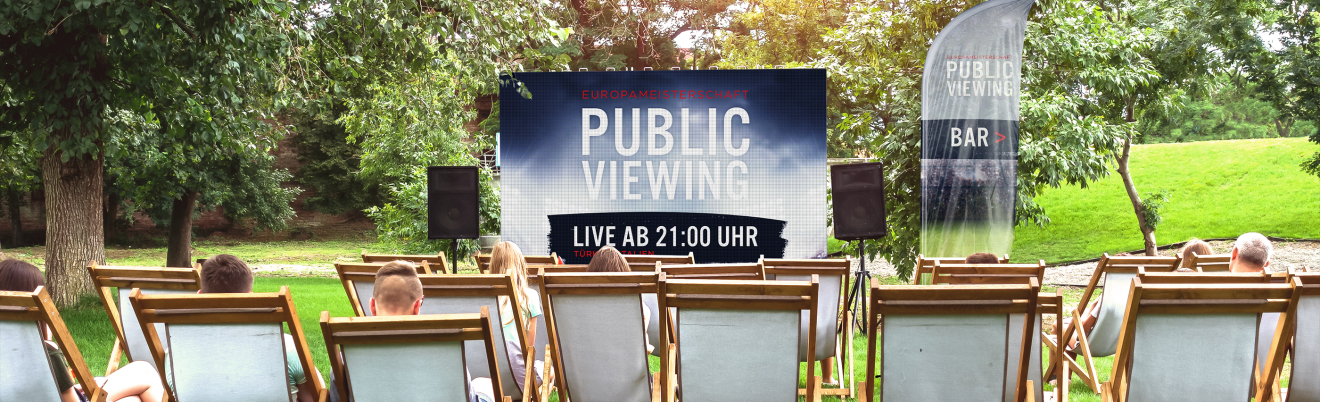 Menschen sitzen auf Liegestühlen im Freien vor einem großen Bildschirm mit der Aufschrift Public Viewing, Live ab 21:00 Uhr in einer Parkanlage mit Bäumen und Gras. Eine Fahne in der Nähe wirbt für eine Bar.