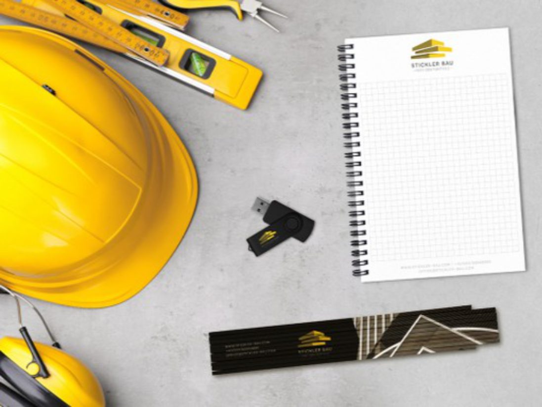 Gelber Bauhelm, Maßband, Wasserwaage, Gehörschutz, USB-Stick, Markenzollstock und ein Spiralnotizblock - diese praktischen Werbemittel sind übersichtlich auf einer hellgrauen Fläche angeordnet.