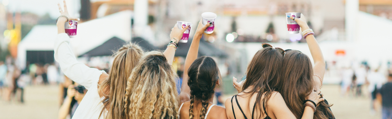 Vier junge Frauen stehen bei einer Veranstaltung im Freien dicht beieinander, mit dem Rücken zur Kamera, heben Becher in die Luft und feiern in einer ausgelassenen, festivalähnlichen Atmosphäre.