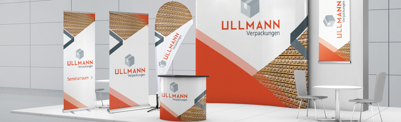 Messestand mit Branding, mit Bannern, einem geschwungenen Hintergrund aus Wellpappe, einer Theke und einem kleinen runden Tisch mit zwei Stühlen in einem modernen Innenraum.